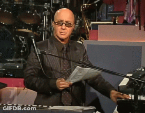 A Bald Man Wearing Glasses Stands In Front Of A Microphone Holding A Piece Of Paper GIF