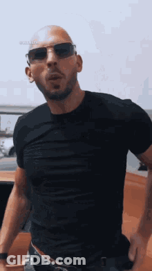 A Bald Man Wearing Sunglasses And A Black Shirt Is Standing In Front Of A Car And Making A Funny Face . GIF