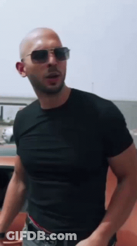 A Bald Man Wearing Sunglasses And A Black T-Shirt Is Standing In Front Of A Car . GIF