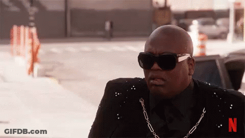 A Bald Man Wearing Sunglasses And A Chain Around His Neck Is Sitting On The Sidewalk . GIF
