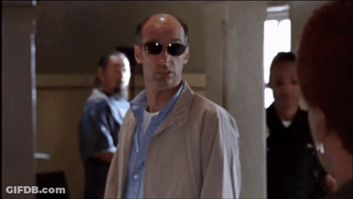 A Bald Man Wearing Sunglasses And A Jacket Stands In A Hallway GIF