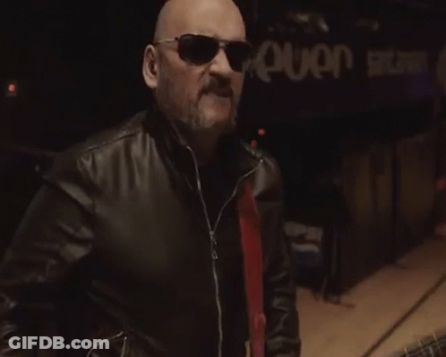 A Bald Man Wearing Sunglasses And A Leather Jacket Is Playing A Guitar GIF