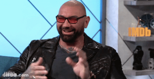 A Bald Man Wearing Sunglasses And A Leather Jacket Is Sitting In Front Of An Imdb Sign GIF