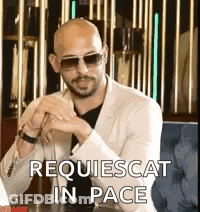 A Bald Man Wearing Sunglasses And A Suit Is Sitting At A Table With His Hands Folded . GIF