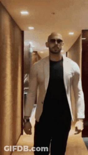 A Bald Man Wearing Sunglasses And A White Jacket Walks Down A Hallway . GIF