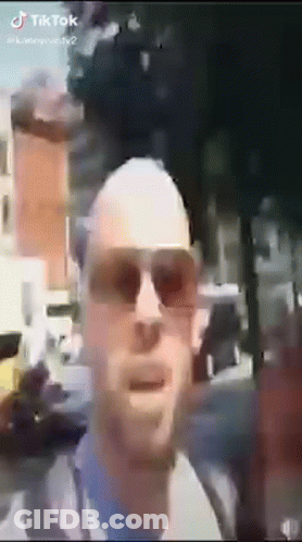 A Bald Man Wearing Sunglasses Is Standing In Front Of A Building . GIF
