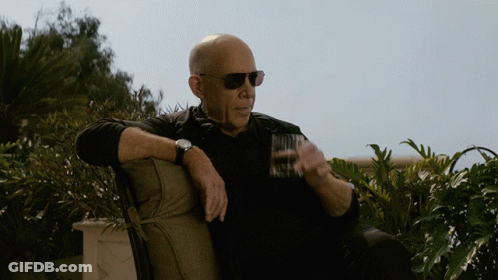 A Bald Man Wearing Sunglasses Sits On A Chair Holding A Glass GIF