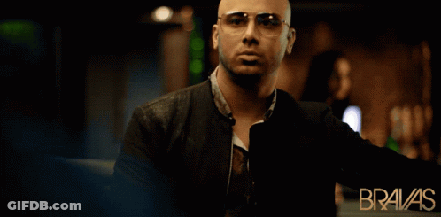 A Bald Man Wearing Sunglasses Stands In Front Of An Ad For Bravas GIF