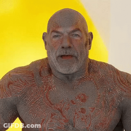 A Bald Man With A Beard And A Tattoo On His Chest Looks At The Camera GIF