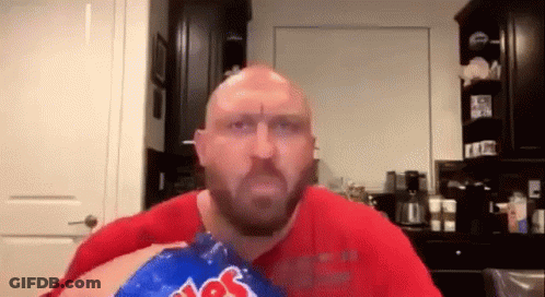 A Bald Man With A Beard Is Eating A Bag Of Potato Chips In A Kitchen . GIF
