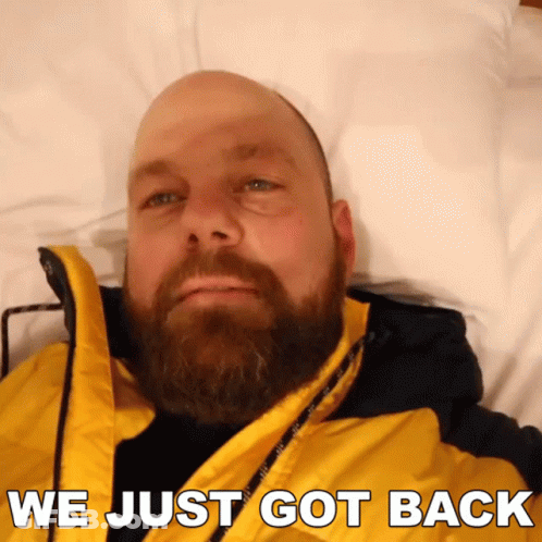A Bald Man With A Beard Is Laying On A Bed With The Words " We Just Got Back " Above Him GIF