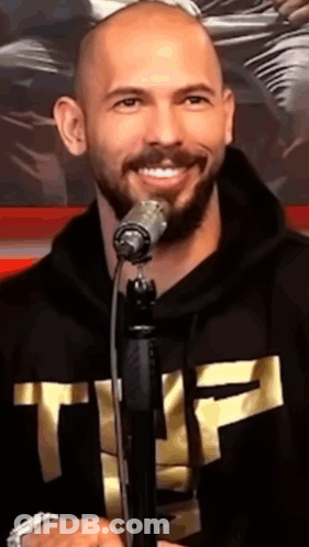 A Bald Man With A Beard Is Singing Into A Microphone While Wearing A Black Hoodie With The Letter T On It GIF