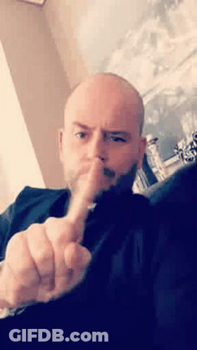 A Bald Man With A Beard Is Sitting On A Couch Making A Rude Gesture With His Middle Finger . GIF