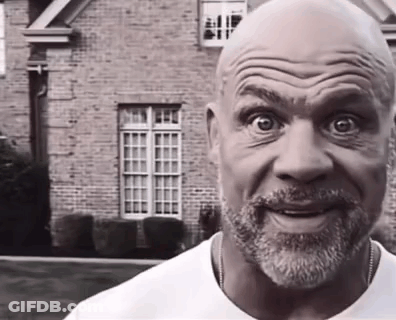 A Bald Man With A Beard Is Smiling In Front Of A Brick Building . GIF