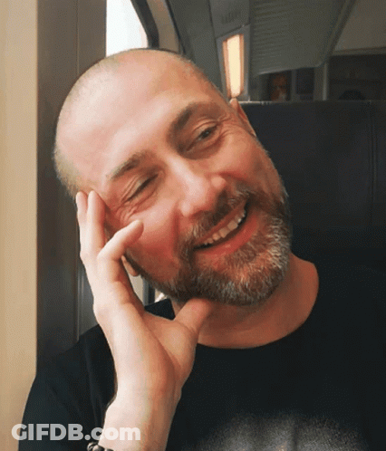 A Bald Man With A Beard Is Smiling With His Hand On His Face GIF