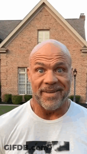 A Bald Man With A Beard Is Standing In Front Of A Brick House And Making A Funny Face GIF