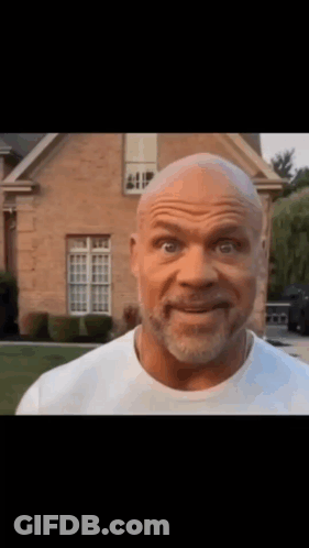 A Bald Man With A Beard Is Standing In Front Of A Brick House . GIF