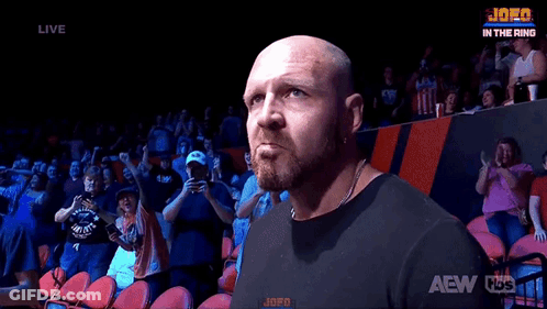 A Bald Man With A Beard Is Standing In Front Of A Crowd In A Stadium . GIF
