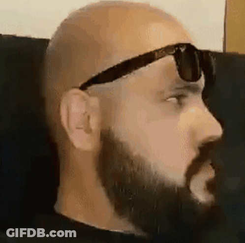 A Bald Man With A Beard Is Wearing Sunglasses While Sitting On A Couch . GIF
