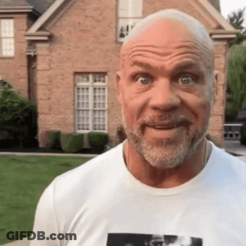 A Bald Man With A Beard Wearing A White T-Shirt Is Standing In Front Of A Brick House GIF