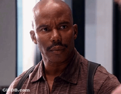 A Bald Man With A Mustache Wearing A Brown Shirt And Suspenders Looks At The Camera . GIF
