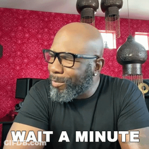 A Bald Man With Glasses And A Beard Is Sitting In Front Of A Sign That Says Wait A Minute GIF
