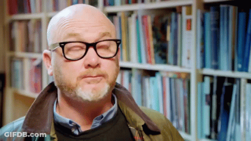 A Bald Man With Glasses And A Beard Is Standing In Front Of A Bookshelf With His Eyes Closed GIF