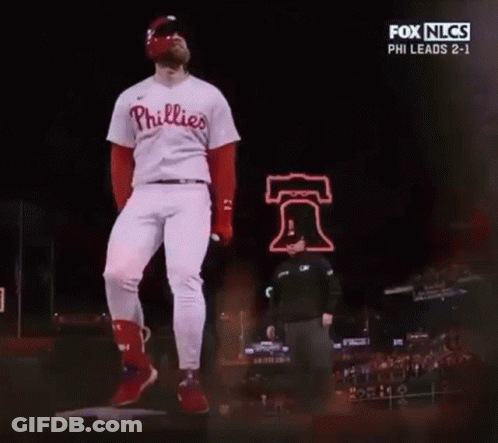 A Baseball Player In A Phillies Uniform Is Standing On A Mound In Front Of A Bell Sign . GIF