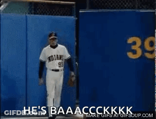 A Baseball Player Is Standing In Front Of A Blue Wall With The Words `` He 'S Baaacckkk '' Written On It . GIF