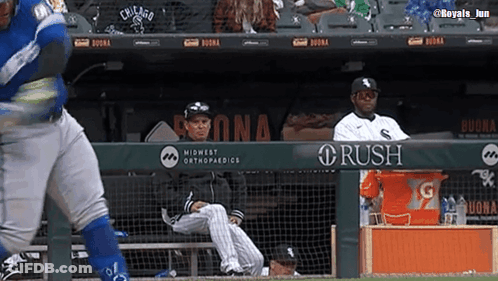 A Baseball Player Is Swinging At A Ball While A Man Sits In The Dugout Watching . GIF