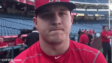 A Baseball Player Wearing A Red Shirt And A Hat Is Standing In A Dugout . GIF