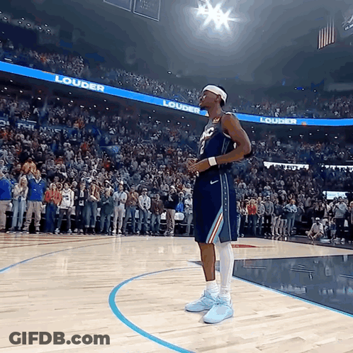 A Basketball Player Stands On The Court In Front Of A Crowd With A Banner That Says Louder On It GIF