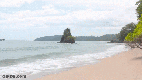 A Beach With A Large Rock In The Middle Of The Water GIF