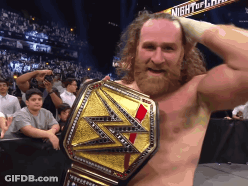 A Bearded Wrestler Is Holding A Wrestling Championship Belt In Front Of A Night Of Champions Sign GIF