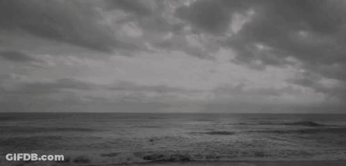 A Black And White Photo Of A Beach With A Cloudy Sky And Waves Crashing On The Shore . GIF