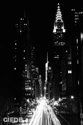 A Black And White Photo Of A City Street At Night With A Tall Building In The Background GIF