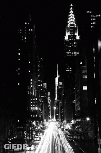 A Black And White Photo Of A City Street At Night With A Tall Building In The Background GIF