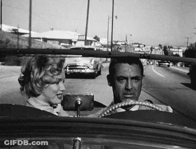 A Black And White Photo Of A Man And A Woman Driving A Car Down A Street . GIF