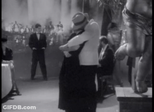A Black And White Photo Of A Man And Woman Hugging In Front Of A Crowd GIF