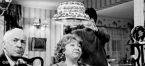 A Black And White Photo Of A Man Holding A Cake Over A Woman 'S Head In A Living Room . GIF