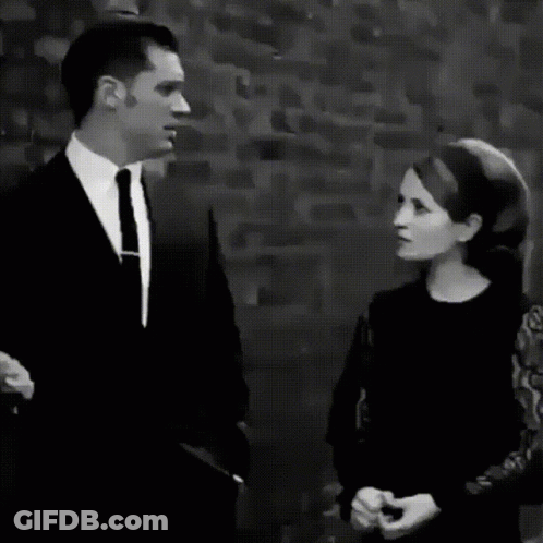 A Black And White Photo Of A Man In A Suit And Tie Standing In Front Of A Brick Wall GIF