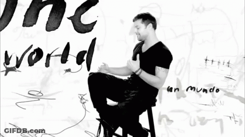 A Black And White Photo Of A Man Sitting In Front Of A Wall With The Word World Written On It GIF