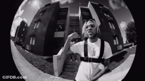 A Black And White Photo Of A Man Standing In Front Of A Building Wearing Sunglasses . GIF