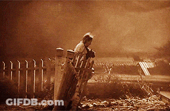 A Black And White Photo Of A Man Standing In Front Of A Fence Holding A Dog GIF
