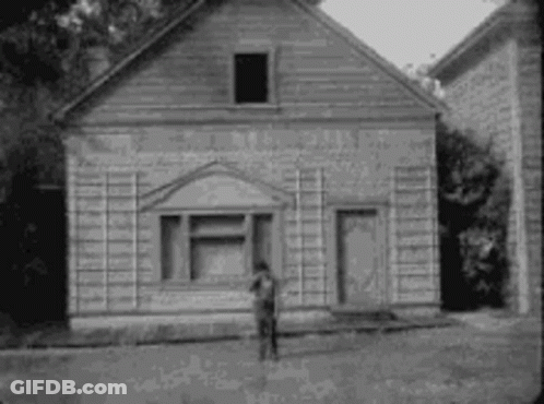 A Black And White Photo Of A Man Standing In Front Of A Small Wooden Building . GIF