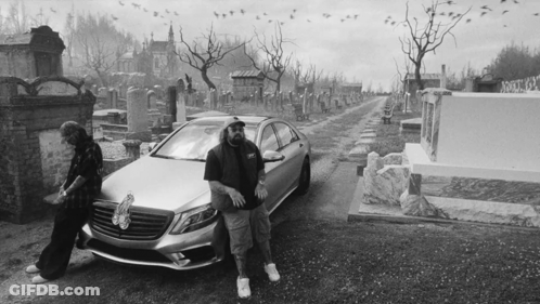 A Black And White Photo Of A Man Standing Next To A Car In A Cemetery GIF
