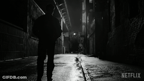 A Black And White Photo Of A Man Walking Down A Dark Alleyway With Netflix Written In The Corner GIF