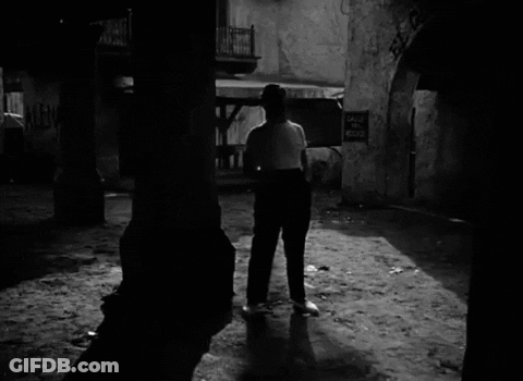 A Black And White Photo Of A Man Walking In Front Of A Sign That Says Calle Del Medio GIF