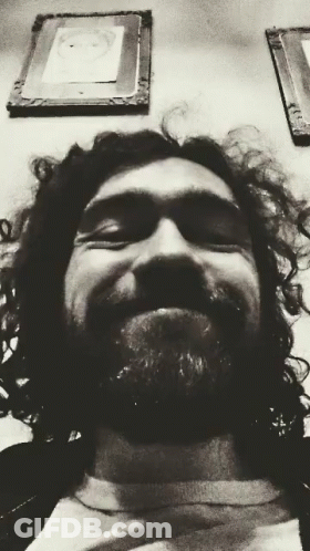 A Black And White Photo Of A Man With Curly Hair And A Beard Smiling GIF