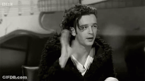 A Black And White Photo Of A Man With Curly Hair Sitting At A Table With His Hand In His Hair . GIF
